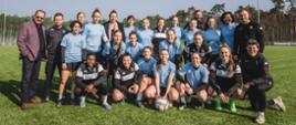 Grupa uśmiechniętych piłkarek Medyka Konin w niebieskich sportowych strojach oraz przedstawiciele klubu i minister Sławomir Nitras pozują do wspólnego zdjęcia na zielonej murawie. 