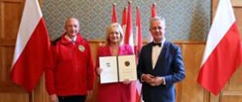 W sali z wyłożonymi drewnianymi panelami ścianami stoi mężczyzna w garniturze, kobieta w różowej marynarce trzymająca otwartą teczkę i mężczyzna w czerwonej kurtce, za nimi pod ścianą polskie flagi.