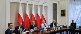 Na zdjęciu kilka osób siedzących za stołem przy mikrofonach, w tle pięć polskich flag.