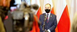 Minister Adam Andruszkiewicz podczas konferencji prasowej, stoi przed mikrofonem, na tle polskich flag.