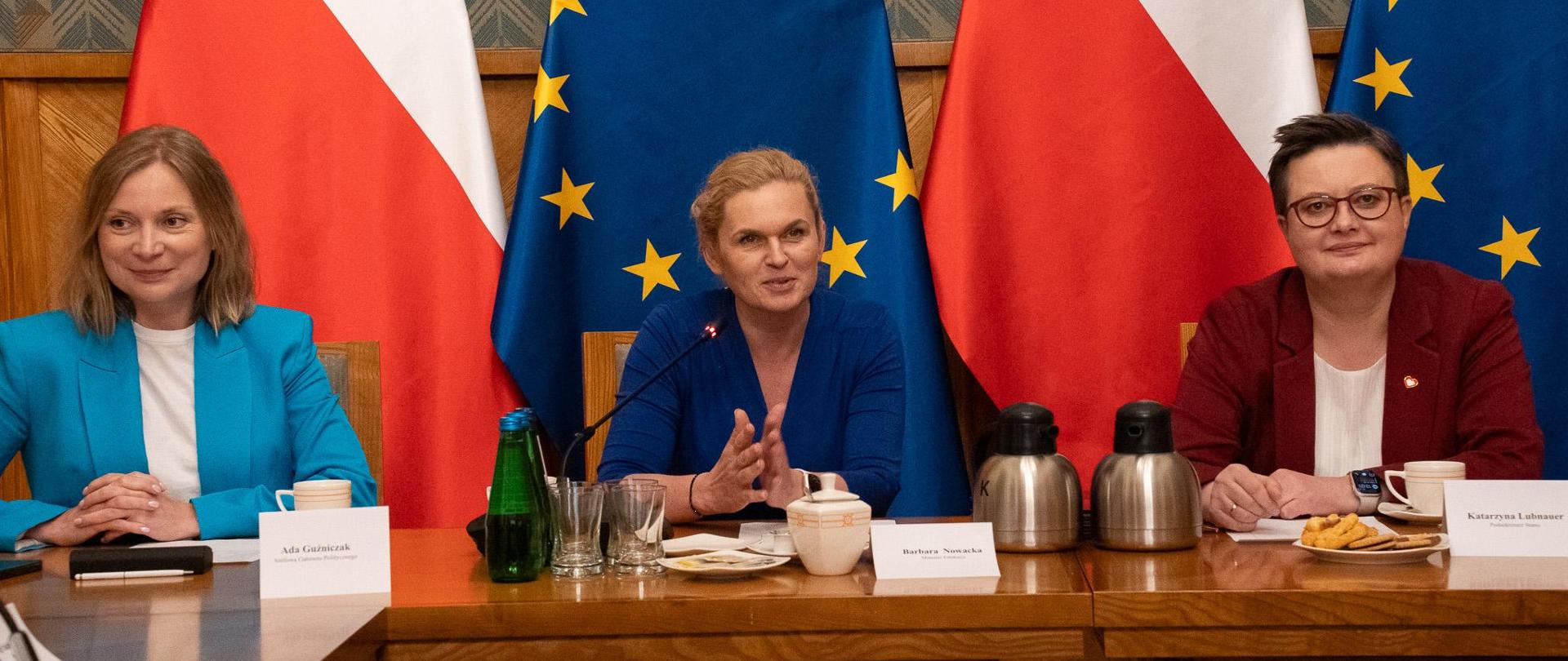 Duża sala ze ścianami w zielony wzorek, przy drewnianym stole siedzi od lewej: Szefowa Gabinetu Politycznego Ada Guźniczak, minister Barbara Nowacki i wiceminister Katarzyna Lubnauer. Na stole rozłożone notatki oraz ustawione napoje. W tle flagi Polski i UE