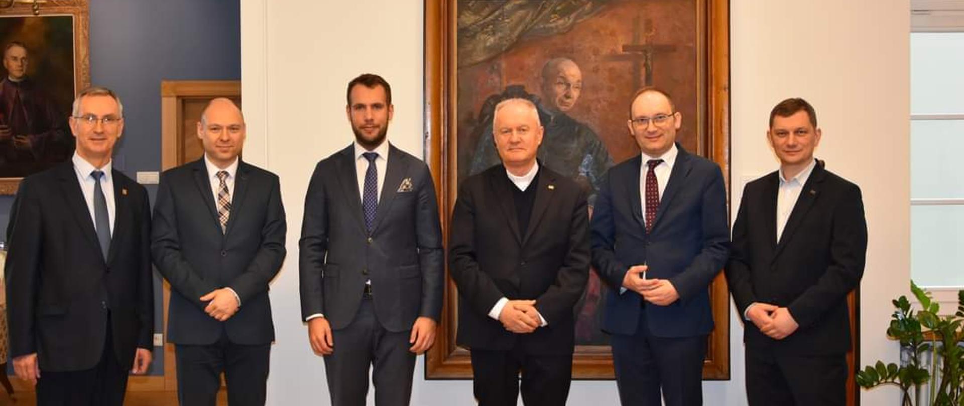 Sześciu mężczyzn w garniturach stoi na tle żółtej ściany, na której wisi duży portret.
