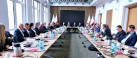Widok z tyłu na wielką salę, przy ustawionych w długi czworokąt stołach siedzą ludzie, z lewej kilka dużych okien, z prawej biała sciana z drewnianymi drzwiami.