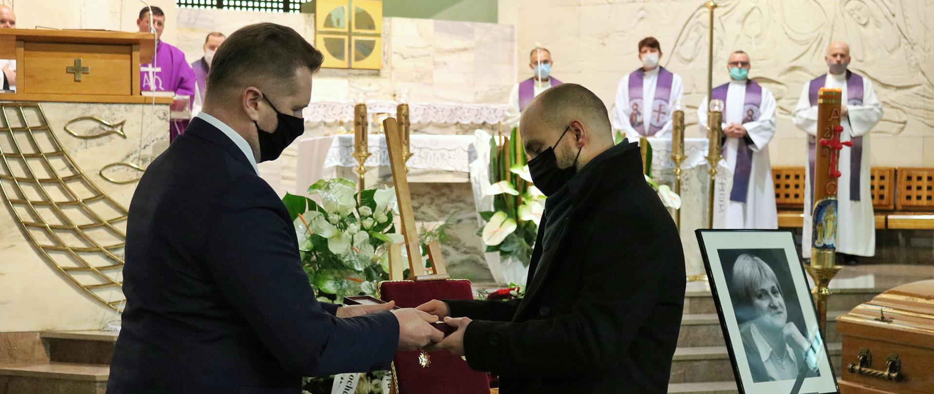 Ceremonia pogrzebowa w kościele. W tle, na ołtarzu duchowni sprawujący liturgię w charakterystycznych w fioletowym kolorze. Przy ołtarzu białe kwiaty, sztalugi z odznaczeniami oraz oraz zdjęcie zmarłej z kirem. Na pierwszym planie minister Przemysław Czarnek wręczający order od Prezydenta RP mężczyźnie z rodziny zmarłej. Obaj panowie w maseczkach na twarzach 