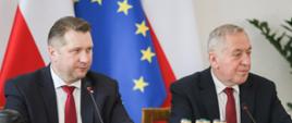 Minister Czarnek i mężczyzna w garniturze siedzą za stołem, przed nimi kilka zielonych butelek z wodą, mikrofony, za nimi flagi Polski i UE.