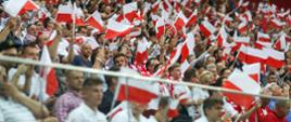 Widok na wypełnione machającymi biało-czerwonymi chorągiewkami ludźmi trybuny.