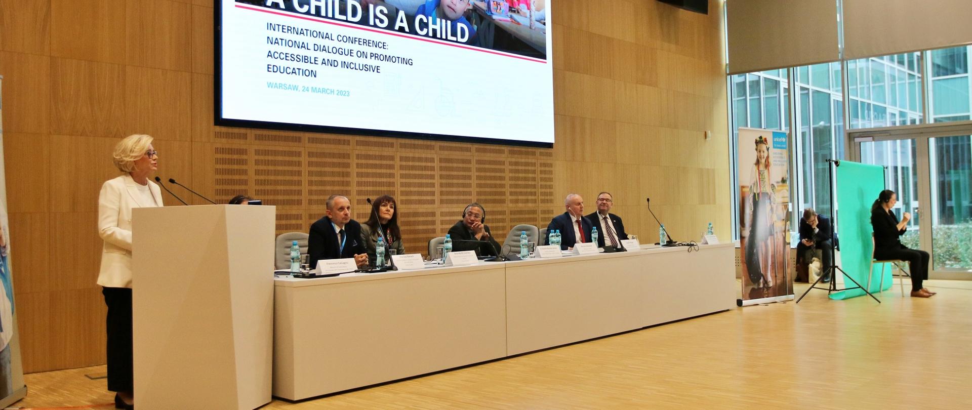 Widok z boku na salę, na drewnianej podłodze stoi biały stół za którym siedzi pięć osób, nad nimi na brązowej ścianie wielki ekran z napisem A child is a child, obok stołu biała mównica za którą stoi wiceminister Machałek i mówi do dwóch mikrofonów.