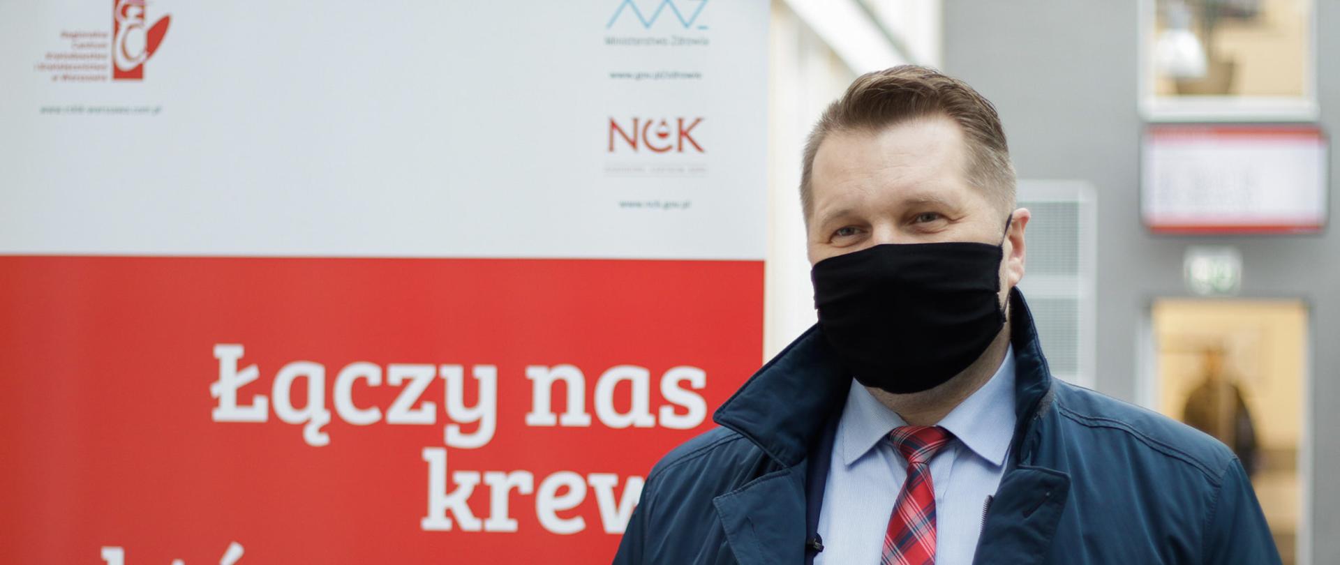 Na zdjęciu minister Przemysław Czarnek ubrany w płaszcz i czerwony krawat w kratce, na twarzy czarna maseczka. Minister stoi na tle czerwonego baneru z napisem łączy nas krew, która ratuje życie. Zdjęcie wykonane w regionalnym centrum krwiodawstwa i krwiolecznictwa w Warszawie. 