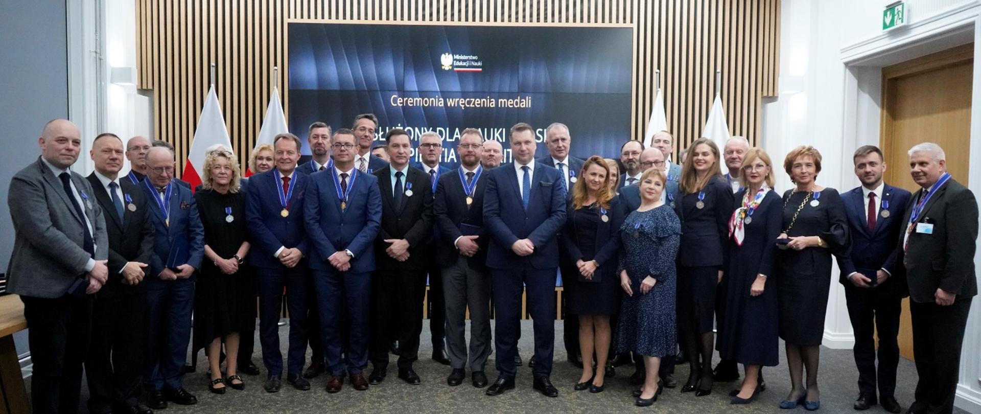 W dużej sali pod ścianą z wielkim ekranem stoi duża grupa osób.