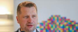 Minister Czarnek mówi do mikrofonu, za nim widać baner z kolorowym geometrycznym wzorem na białym tle.