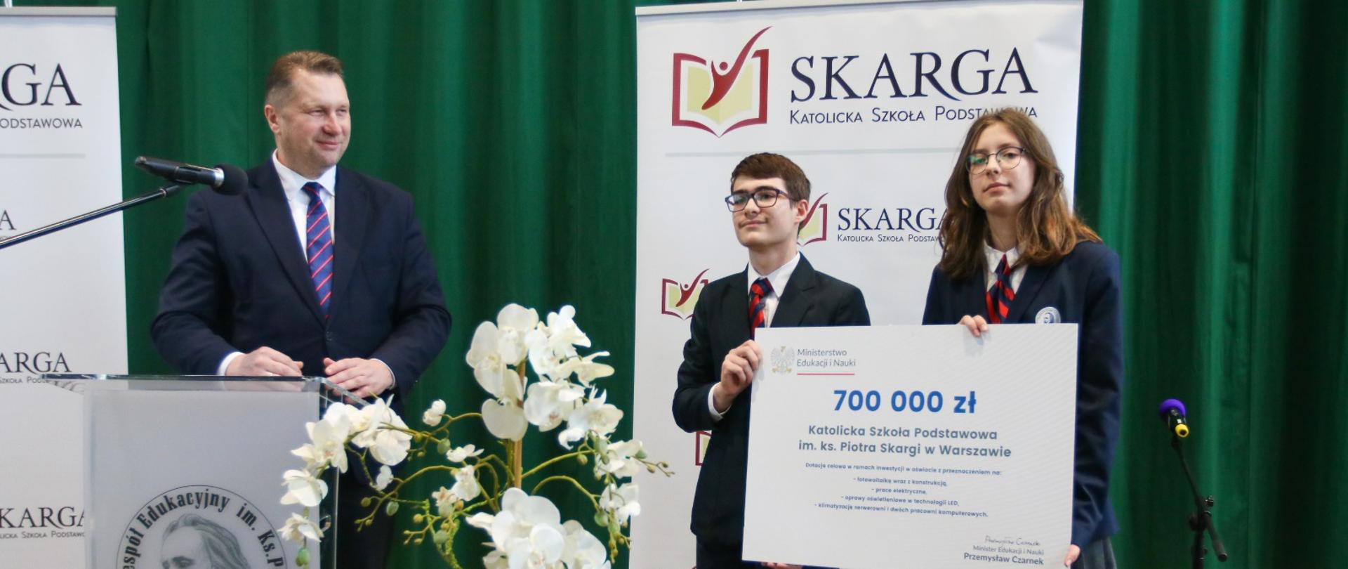 Minister Czarnek stoi za mównicą, patrzy na stojącą obok parę młodych elegancko ubranych ludzi trzymających wielki symboliczny czek z napisem 700 000 zł, obok donica z kwitnącą na biało rośliną, dalej ścianka z napisem Skarga, w tle ciemnozielona kotara.