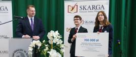 Minister Czarnek stoi za mównicą, patrzy na stojącą obok parę młodych elegancko ubranych ludzi trzymających wielki symboliczny czek z napisem 700 000 zł, obok donica z kwitnącą na biało rośliną, dalej ścianka z napisem Skarga, w tle ciemnozielona kotara.