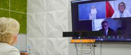 Wiceminister Machałek w białym ubraniu siedzi przy jasnoszarym biurku, przed nią duży monitor na którym widać innych uczestników spotkania.