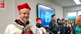 Uśmiechnięty mężczyzna w czerwonym ceremonialnym stroju z białą futrzaną narzutką i w czerwonej czapce stoi za mównicą i mówi do mikrofonu, w reku trzyma metalową buławę, za nim stoi kobieta w takim samym stroju, dalej szereg osób w czarnych togach i czarnych czapkach, za nimi na ścianie duży monitor z napisem 30 lat, w tle okno.
