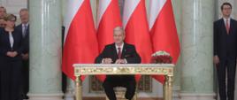 Jerzy Kwieciński podczas uroczystości nominacji na ministra finansów, inwestycji i rozwoju, siedzi za stołem z długopisem w ręku, w tle polskie flagi