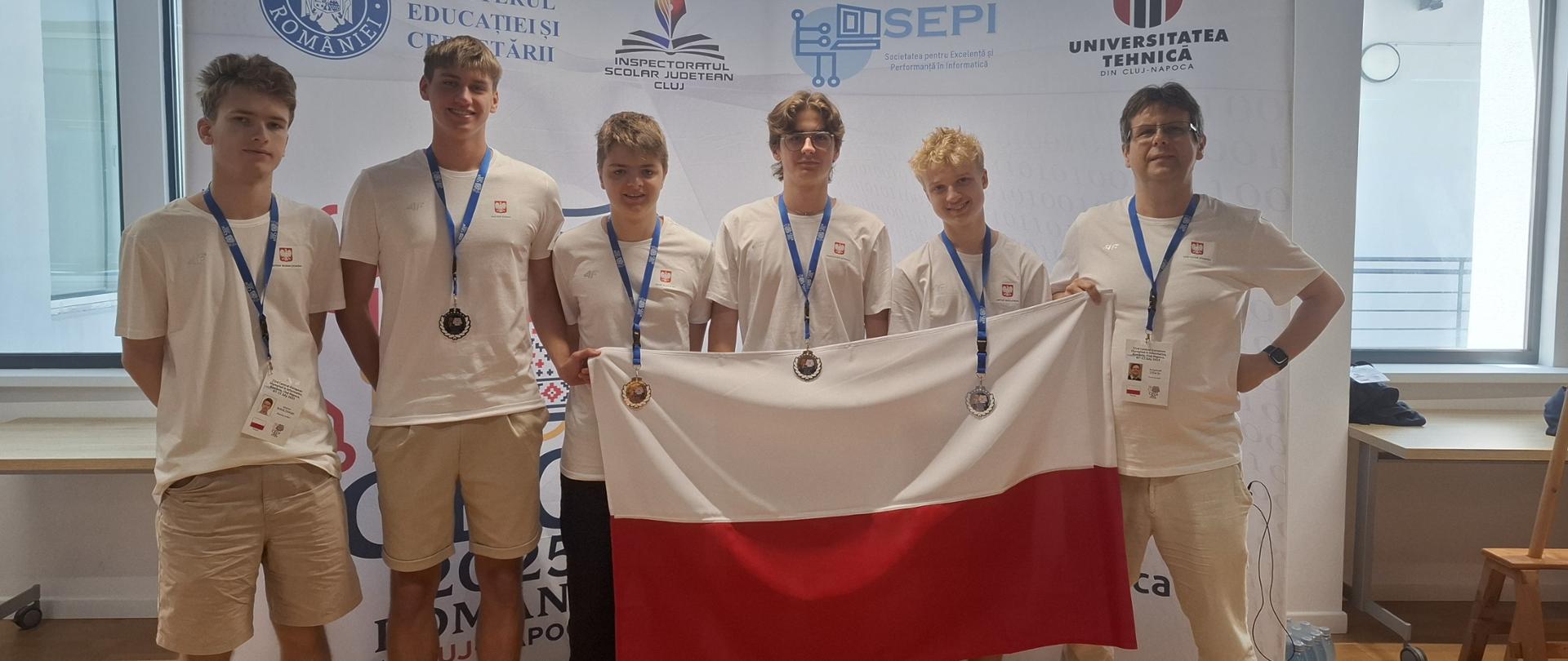 Zgromadzenie sześciu osób, czterech młodych mężczyzn i dwóch dorosłych mężczyzn, trzymających polską flagę. Wszyscy ubrani są w białe koszulki i mają medale zawieszone na szyjach. W tle widać baner z napisem „GUVERNUL ROMANIEI MINISTERUL EDUCATIEI”