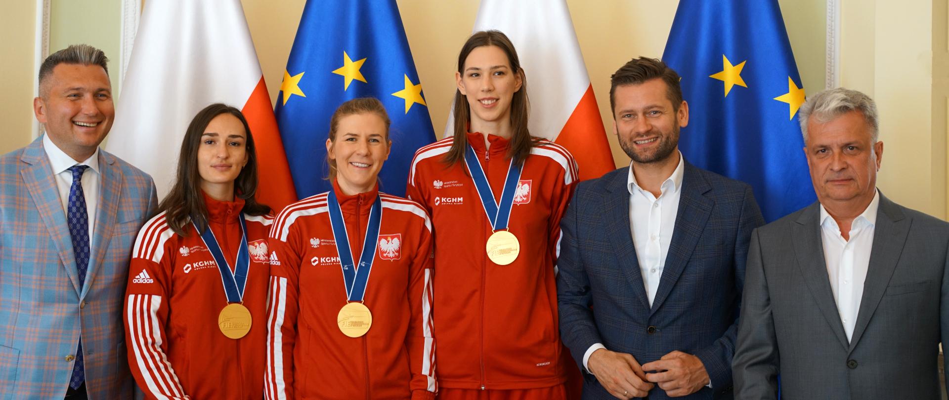 Minister Sportu i Turystyki Kamil Bortniczuk stoi razem z trzema szpadzistkami, które jako drużyna zdobyły mistrzostwo świata. Zawodniczki mają złote medale i są ubrane w czerwone dresy sportowe z polskim godłem. Szpadzistki w składzie: Martyna Swatowska-Wenglarczyk, Ewa Trzebińska i Magdalena Pawłowska stoją razem z ministrem Kamilem Błotniczkiem, prezesem Komitetu Olimpijskiego Radosławem Piesiewiczem i przedstawicielem Polskiego Związku Szermierczego na tle stojących flag Polski i Unii Europejskiej.