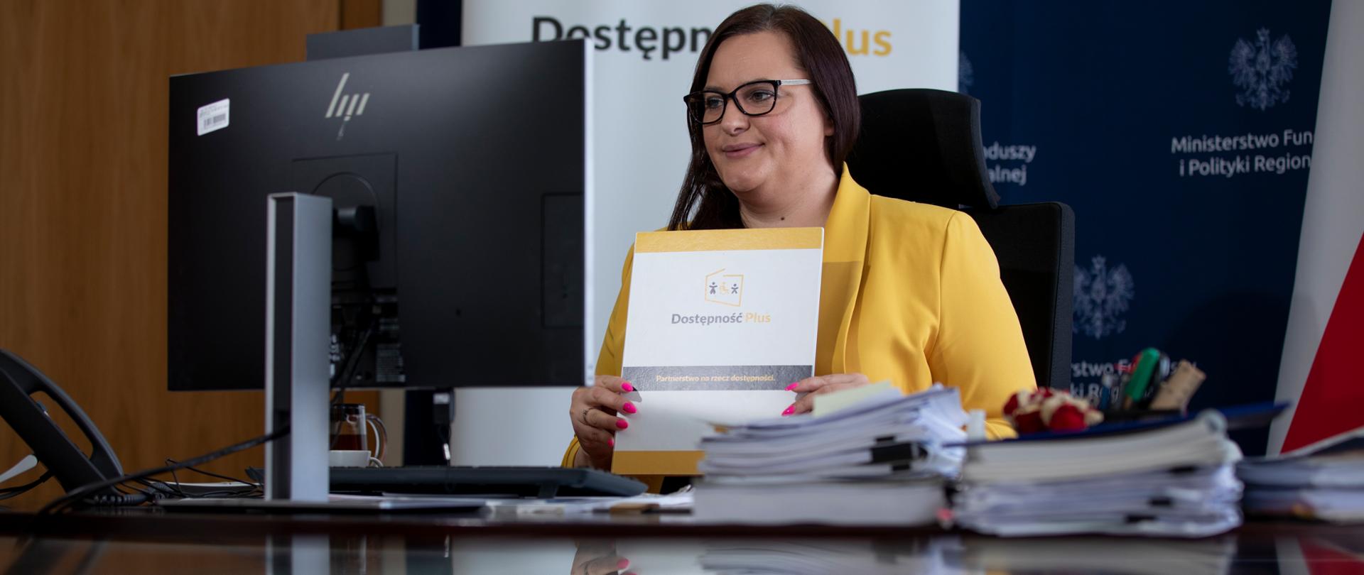 Wiceminister Jarosińska-Jedynak siedzi prze monitorem i trzyma w ręku dokument Dostępność Plus. Za nią ścianka ministerialna, dwie flagi PL i UE oraz rollup programu D+.