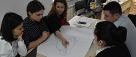 Cztery kobiety i mężczyzna pochylają się nad kartką papieru, na której narysowany jest diagram.
