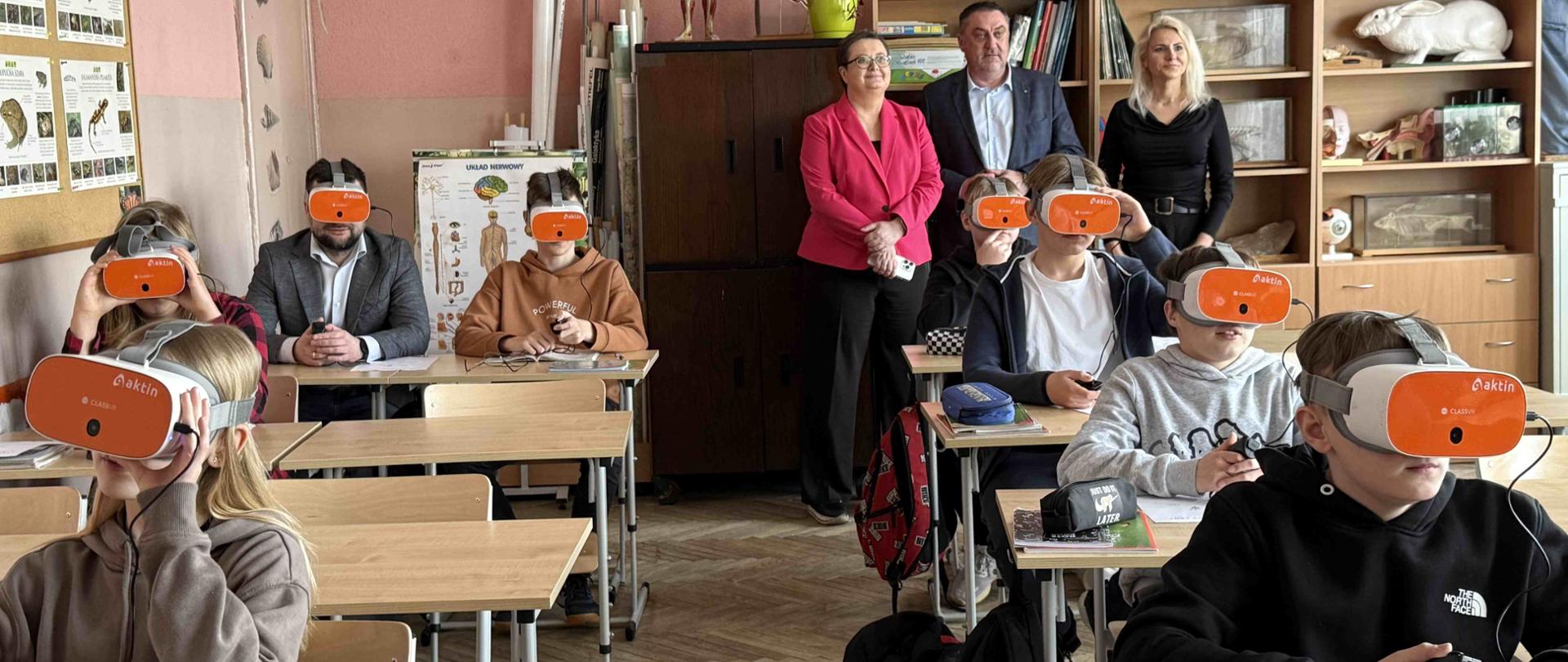 Widok z przodu na klasę szkolną, gdzie w ławkach siedzą dzieci z okularami VR.Z tyłu stoi trójka dorosłych.
