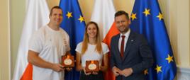 Minister sportu i turystyki Kamil Bortniczuk stoi wraz z srebrnymi medalistami mistrzostw świata w lekkiej atletyce, Natalią Kaczmarek i Wojciechem Nowickim. Zawodnicy prezentują przed sobą zdobyte medale, a tło stanowią flagi Polski i Unii Europejskiej. 