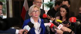 Wiceminister Machałek w niebieskim ubraniu stoi i mówi, dookoła niej dziennikarze trzymający mikrofony.
