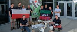 Przed wejściem do budynku stoi grupa młodych ludzi, trzymają polską flagą i zielono-czarno-czerwoną flagę z napisem ADH, przed nimi mały robot na czterokołowym podwoziu.
