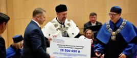 Minister Czarnek stoi obok mężczyzny w ceremonialnym stroju z gronostajowym kołnierzem, razem trzymają wielki symboliczny czek z napisem 25 500 000 zł, obok nich stoi mężczyzna w czarno-niebieskim stroju.