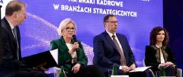Za tle fioletowej ściany z napisem Zawodowcy potrzebni od zaraz siedzą cztery osoby, dwóch mężczyzn w garniturach, kobieta ubrana na czarno i wiceminister Machałek w zielonym ubraniu, która mówi do trzymanego w ręku mikrofonu.