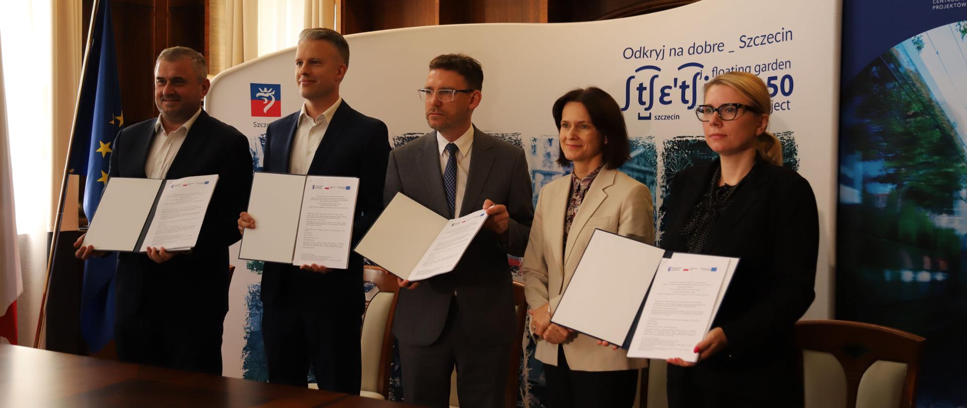 Pięć osób stoi w szeregu i prezentuje dokumenty. Pierwsza z prawej wiceministra Monika Sikora.