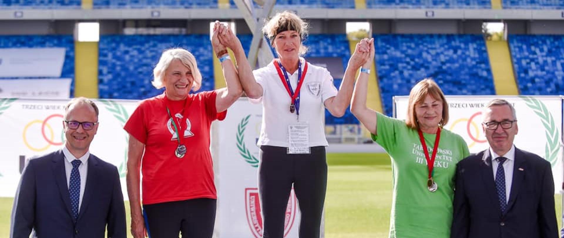 Wiceminister Stanisław Szwed na Stadionie Śląskim w Chorzowie obok stojących na podium trzech medalistek XII Międzynarodowej Olimpiady Sportowej Uniwersytetów Trzeciego Wieku i Organizacji Senioralnych „Trzeci Wiek na Start”.
