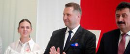 Minister Czarnek, mężczyzna w garniturze i dziewczyna w białej bluzce stoją w rzędzie, minister bije brawo.