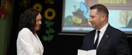 Minister Czarnek stoi i podaje rękę ubranej na biało kobiecie, w drugiej ręce trzyma kartkę papieru.