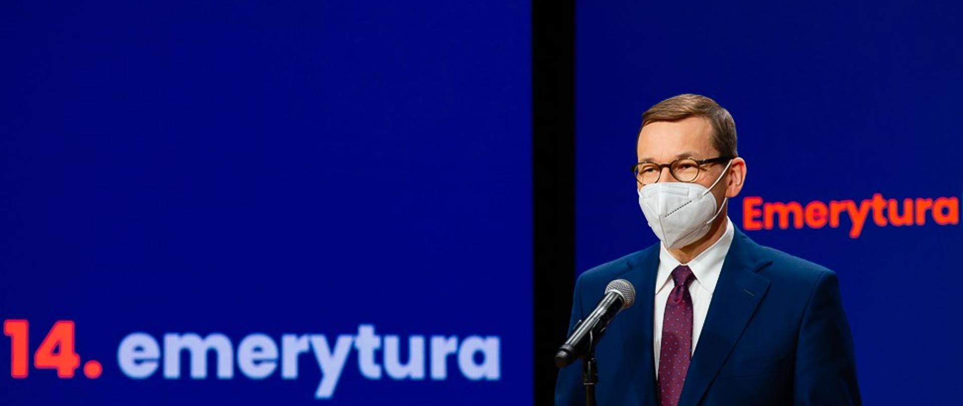 Premier Mateusz Morawiecki stoi przy mikrofonie. W tle ścianka promocyjna z napisem "14. emerytura".
