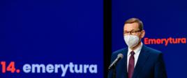 Premier Mateusz Morawiecki stoi przy mikrofonie. W tle ścianka promocyjna z napisem "14. emerytura".