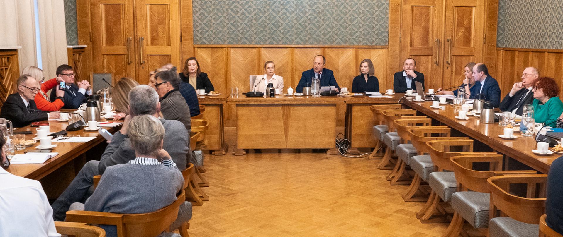 Grupa kobiet i mężczyzn siedzi przy stole ustawionym w literę U. Na stole stają filiżanki, dzbanki i mikrofony. 