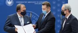W tle baner Uniwersytetu Medycznego w Lublinie. Minister edukacji i nauki Przemysław Czarnek przekazuje rektorowi Uniwersytetu Medycznego w Lublinie otwartą teczkę z dokumentami. Po prawej stronie minister zdrowia Adam Niedzielski. Na twarzach mają maseczki. 
