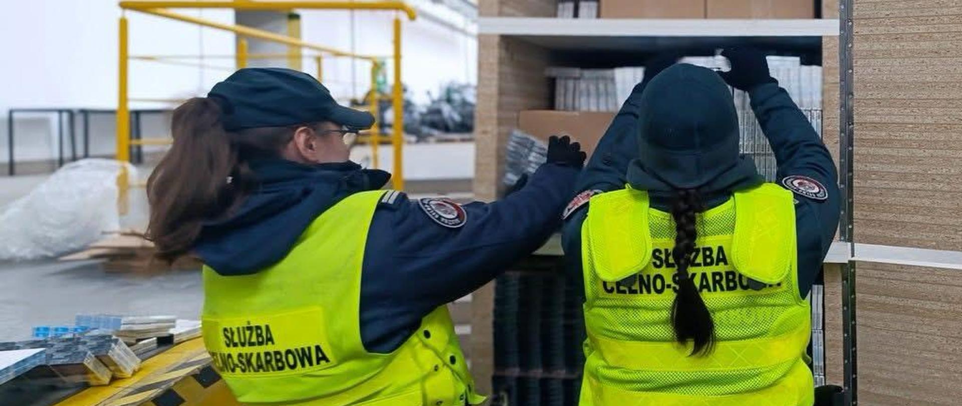 Dwie osoby w kamizelkach z napisem „Służba Celno‑Skarbowa” stoją przy metalowym regale i sprawdzają ułożone na półkach opakowania; w tle widać elementy hali magazynowej oraz żółtą barierkę.
