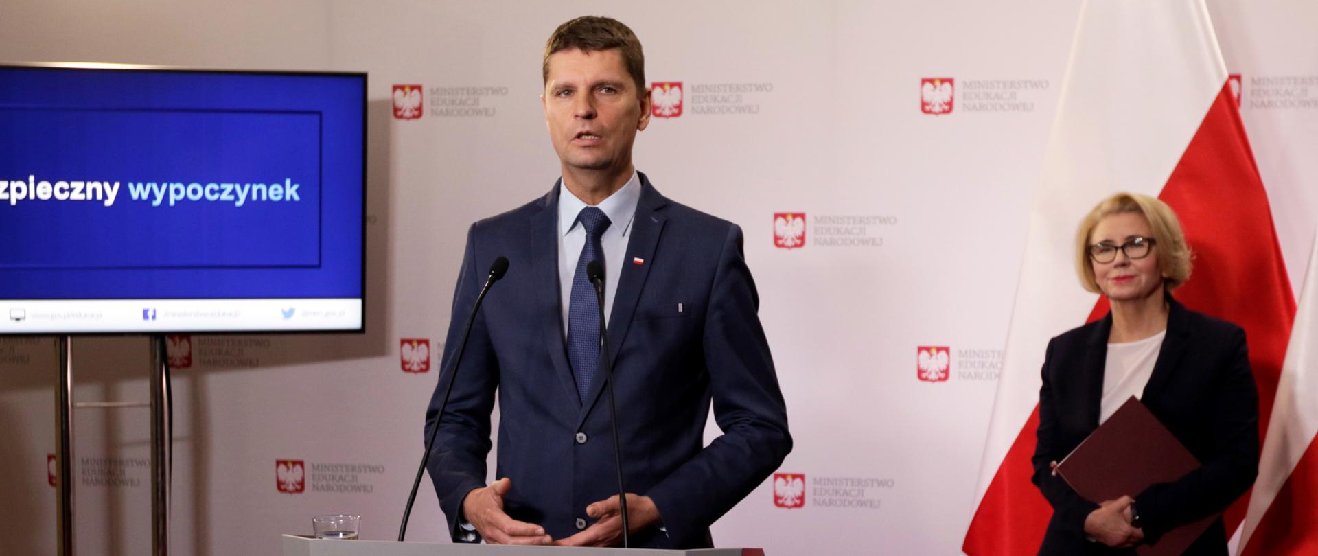 Minister edukacji przemawia na konferencji. Stoi za mównicą, po lewej stronie monitor z prezentacją a po prawej stronie stoi wiceminister edukacji Marzena Machałek. 