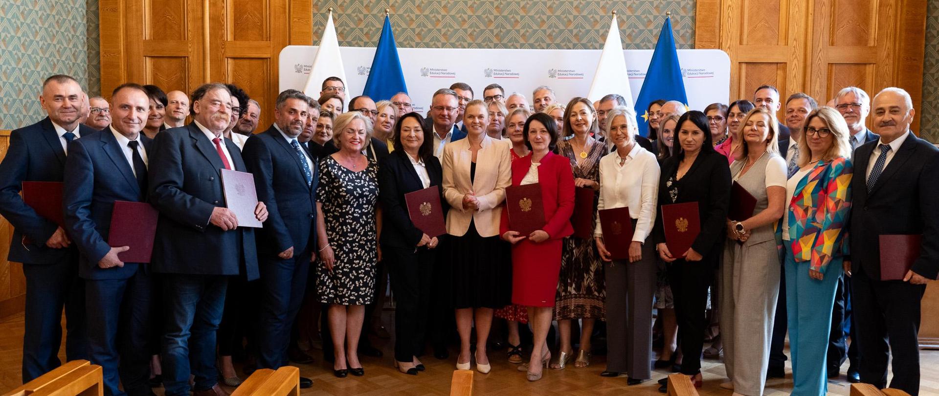 W sali na tle ścianki stoją powołani członkowie Rady Dyrektorów Szkół i Placówek Szkolnictwa Branżowego i pozują do zdjęcia. Większość z nich w ręku trzyma teczki z powołaniem. Pośrodku stoi minister Barbara Nowacka. 