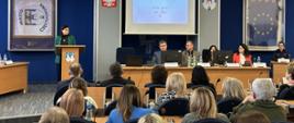 Obraz przedstawia ustawienie sali konferencyjnej z kilkoma osobami siedzącymi i jedną osobą stojącą na podium. Podium znajduje się z przodu sali, nieco po lewej stronie, i zajmuje je kobieta ubrana w zieloną kurtkę. Za nią na ścianie wywieszony jest sztandar z napisem "MIASTO CZĘSTOCHOWA" i herbem.
Na pierwszym planie znajduje się kilka rzędów uczestników siedzących przy biurkach, zwróconych twarzą do podium i ekranu projekcyjnego. Wśród uczestników przeważają kobiety, wśród których przeplata się kilku mężczyzn. Siedzą na krzesłach z zaokrąglonymi oparciami, a niektórzy mają przed sobą zeszyty lub papiery. Na jednym z biurek widoczna jest butelka wody.
Z przodu sali za długim biurkiem siedzi cztery osoby. Każdy z panelistów ma przed sobą mikrofon i tabliczkę z nazwiskiem. Wśród panelistów są dwaj mężczyźni i dwie kobiety. Nad panelistami na ekranie projekcyjnym wyświetlany jest slajd prezentacji.
