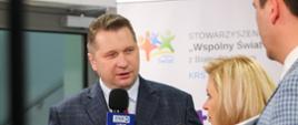 Minister mówi do trzymanego w ręku mikrofonu z napisem TVP3, obok niego stoi jasnowłosa kobieta trzymająca taki sam mikrofon i mężczyzna w marynarce w szarą kratkę.