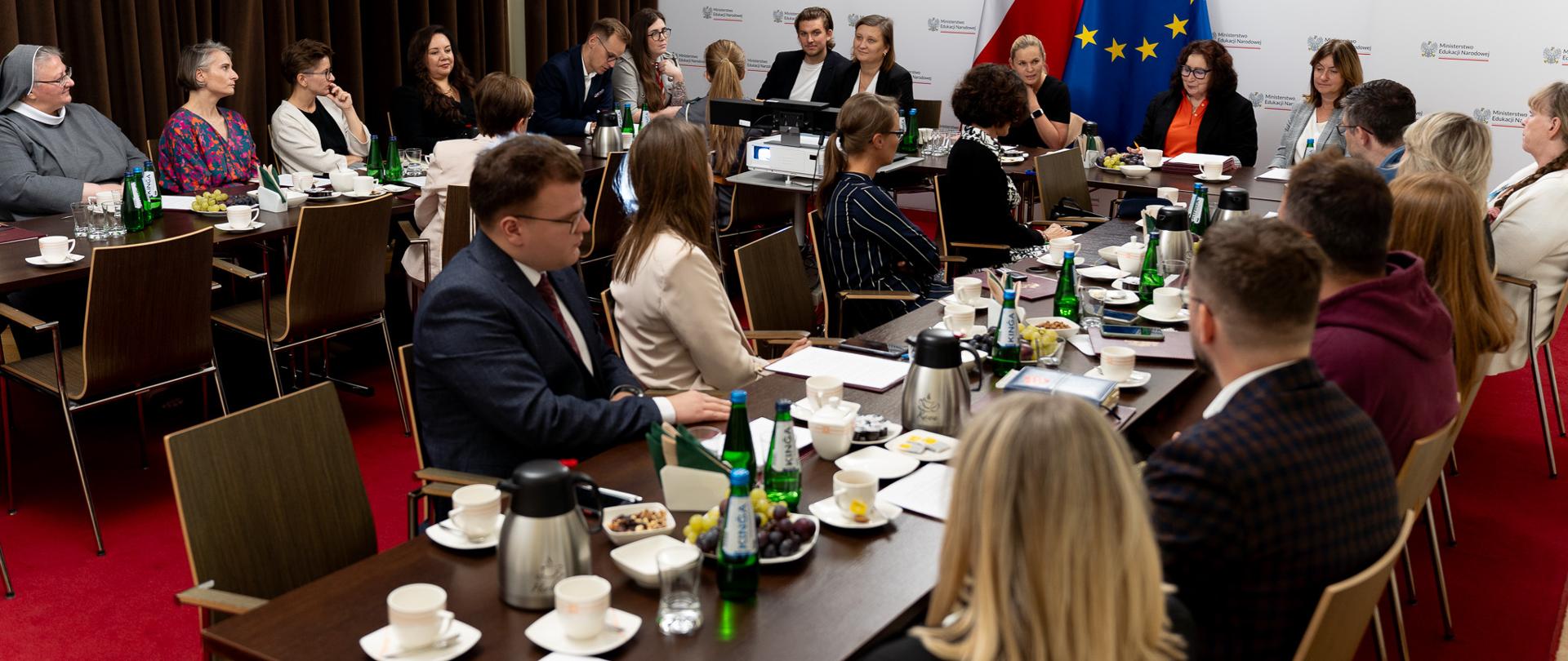Grupa ludzi siedzi wokół dużego prostokątnego stołu w sali konferencyjnej. Stół wypełniony jest różnymi przedmiotami, w tym laptopami, zeszytami, papierami, kubkami do kawy, butelkami z wodą i projektorem.
W tle na tle białej ścianki siedzi minister Nowacka i wiceminister Piechna-Więckieiwcz