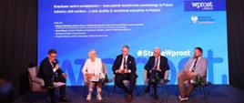 Pod niebieską ścianą na krzesłach siedzi wiceminister Machałek i czterech mężczyzn w garniturach, jeden z nich mówi do trzymanego w ręku mikrofonu.