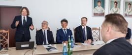 Grupa ludzi siedzi przy stole, jeden mężczyzna stoi i coś mówi, gestykulując rękami.
