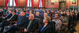 Grupa osób siedzi na konferencji w sali z portretami 