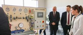 Minister Czarnek, kobieta w białej bluzce i mężczyzna w garniturze stoją obok tablicy rozdzielczej z szeregiem zegarów i przełączników, w tle duże okno.