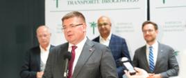 Pod ścianką z napisem Inspekcja Transportu Drogowego stoi trzech mężczyzn w garniturach, przed nimi wiceminister Rzymkowski stoi i mówi do mikrofonu na stojaku.
