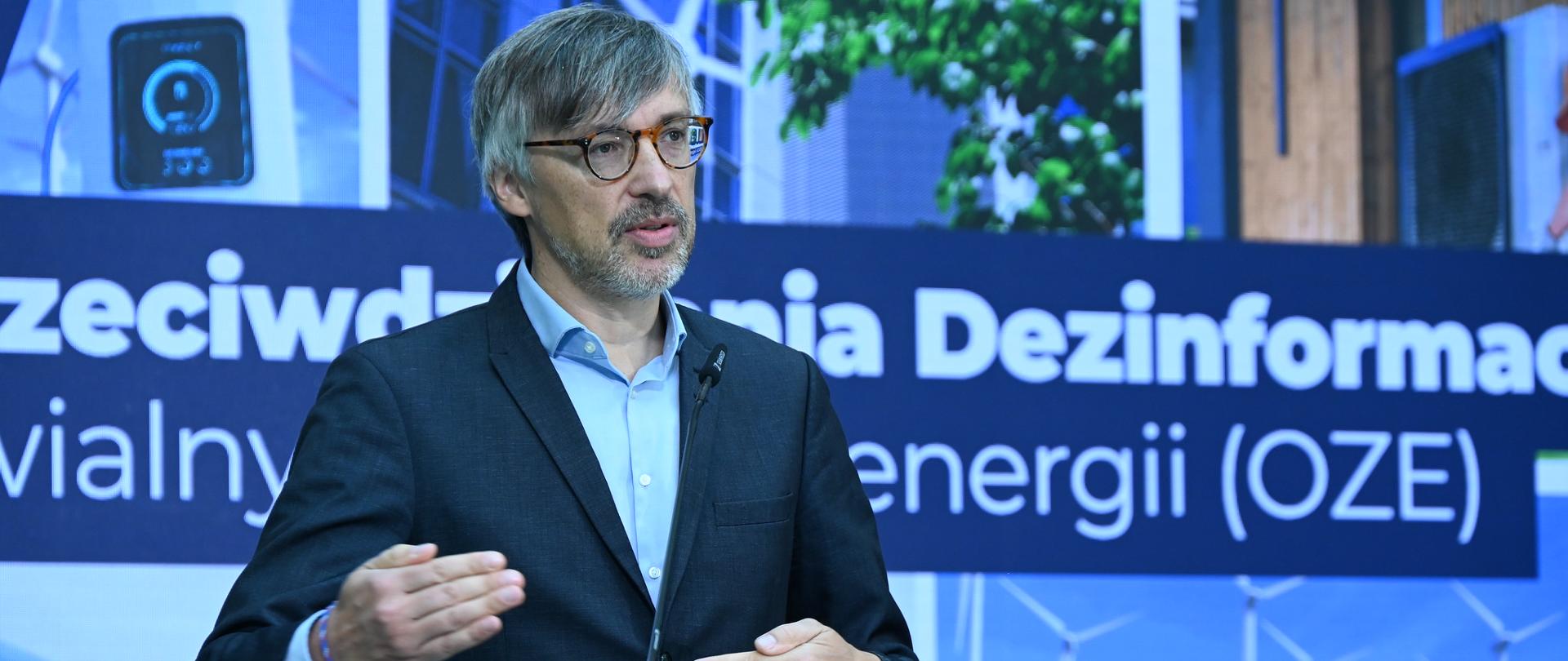Osoba stoi przy przezroczystym podium i przemawia do mikrofonu. W tle widoczna jest plansza poświęcona przeciwdziałaniu dezinformacji dotyczącej odnawialnych źródeł energii. Na planszy znajdują się grafiki przedstawiające instalacje fotowoltaiczne, turbiny wiatrowe, stację ładowania samochodu elektrycznego oraz elementy infrastruktury energetycznej. Osoba wykonuje gest dłonią podczas wystąpienia. Dominują niebieskie barwy.