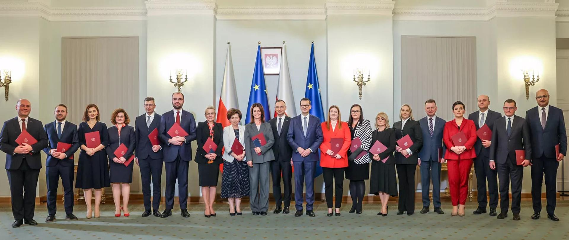 Zdjęcie przedstawia powołaną nową radę ministrów. W centrum stoi Prezydent Andrzej Duda, a bezpośrednio koło niego stoi Premier Mateusz Morawiecki i Danuta Dmowska-Andrzejuk, nowo powołany minister sportu i turystyki. Na zdjęciu znajdują się wszyscy nowo powołani ministrowie w strojach galowych oraz granatowymi teczkami. 